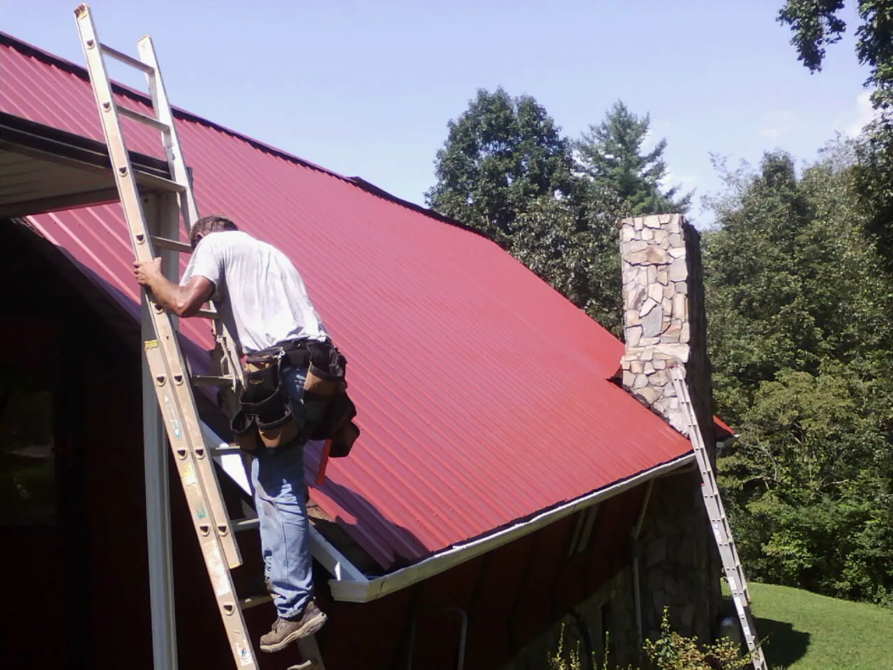 Roofer on a ladder beside a red metal roof and stone chimney — Soffit & Fascia Repair by Roofing Brothers in Maumee